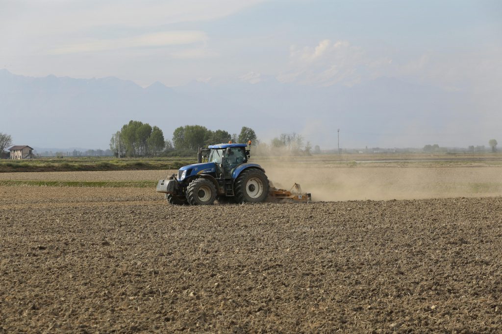 A blue tractor tills an expansive field on a sunny day, creating dust clouds.