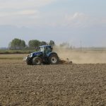 A blue tractor tills an expansive field on a sunny day, creating dust clouds.