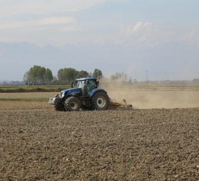 A blue tractor tills an expansive field on a sunny day, creating dust clouds.