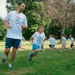 A group of kids running with an instructor during outdoor sports activities on a sunny day in a park.