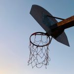 Low-angle view of a worn basketball hoop with a rusted net against a clear blue sky.
