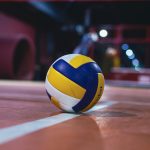 Yellow and white volleyball resting on an indoor volleyball court with bright lights.